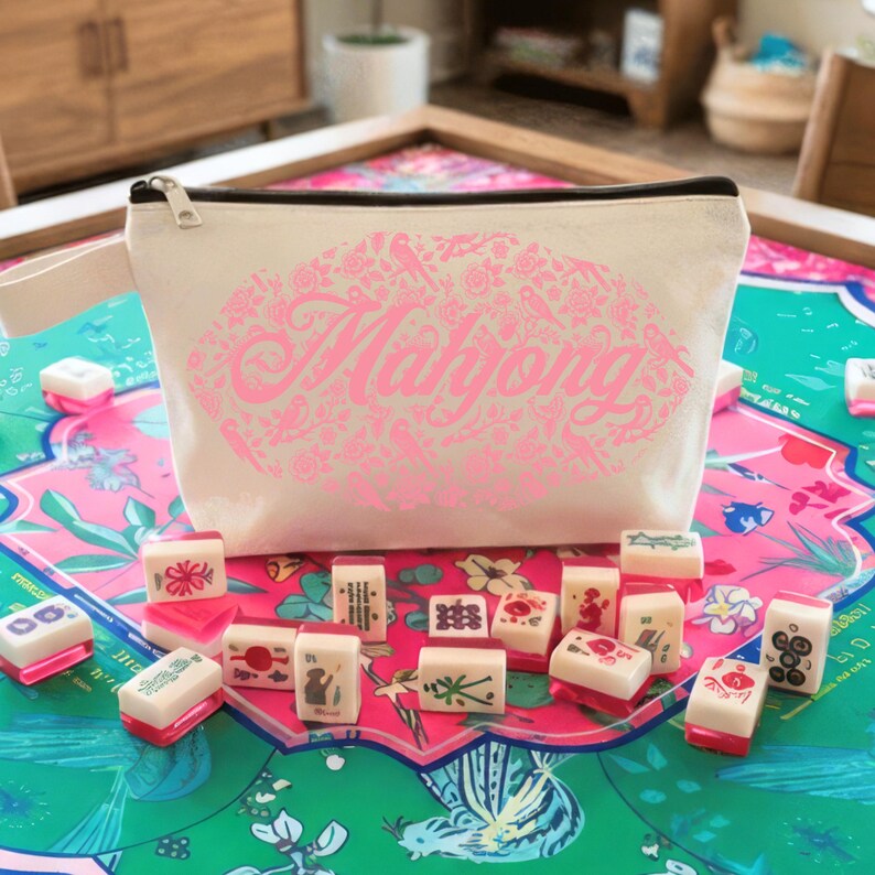May include: A cream-colored zippered pouch with the word "Mahjong" in pink script, surrounded by floral and bird designs. The pouch is next to a set of Mahjong tiles on a colorful game board.