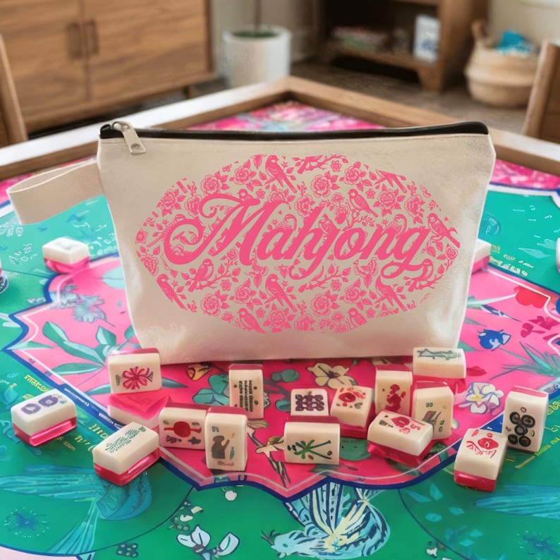 May include: A cream-colored Mahjong cosmetic bag with a black zipper and a pink floral design. The bag features the word "Mahjong" in pink script. Mahjong tiles are scattered on a colorful game board.