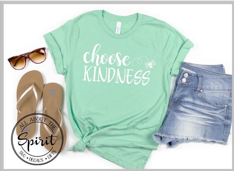 Pu&ograve; includere: Una maglietta verde chiaro con il testo "choose kindness" in lettere bianche. Un piccolo grafico di un'ape &egrave; a destra del testo. La maglietta &egrave; disposta su una superficie bianca con un paio di infradito marroni e un paio di pantaloncini di jeans blu chiaro.