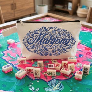 May include: A white canvas Mahjong bag with a blue floral and bird design, and the word "Mahjong" in blue script. The bag is zippered and has a small handle. Mahjong tiles are scattered on a colorful floral game mat.