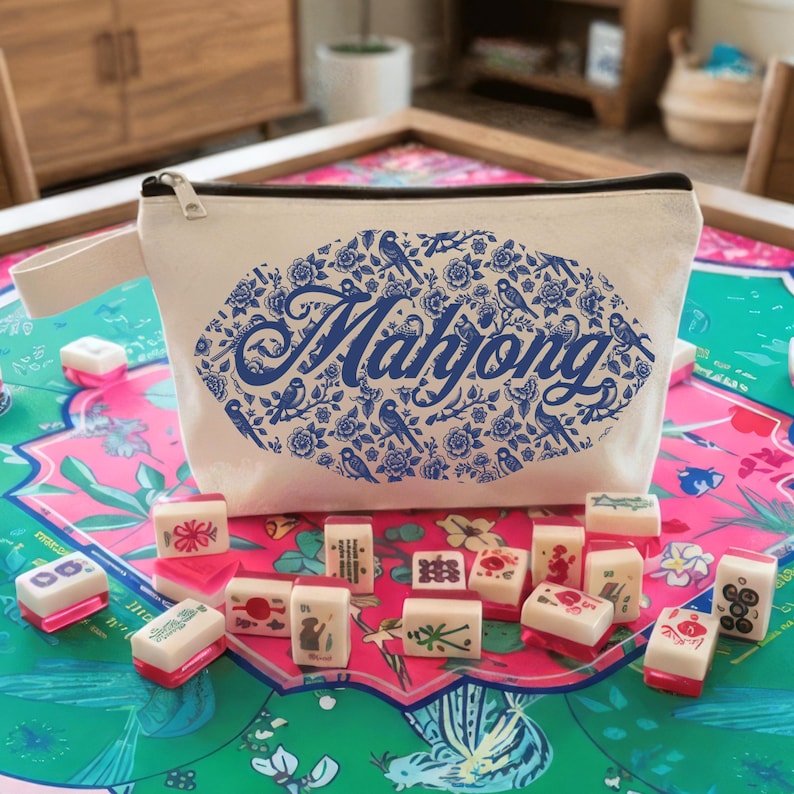 May include: A white canvas Mahjong bag with a blue floral and bird design, and the word "Mahjong" in blue script. The bag is zippered and has a small handle. Mahjong tiles are scattered on a colorful floral game mat.