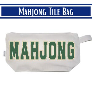May include: A white rectangular zippered pouch featuring the word MAHJONG printed in large, bold, dark green block letters with a thin gold outline. The bag includes a small fabric side tab and a matching white fabric pull loop on the right side.