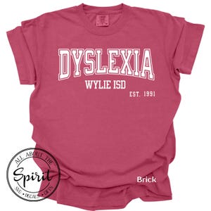 May include: Brick-colored t-shirt with the word "DYSLEXIA" in white, arched text. Below, "WYLIE ISD" and "EST. 1991" are printed. A circular logo with "ALL ABOUT THE Spirit SVG DECALS GIFTS" is at the bottom.