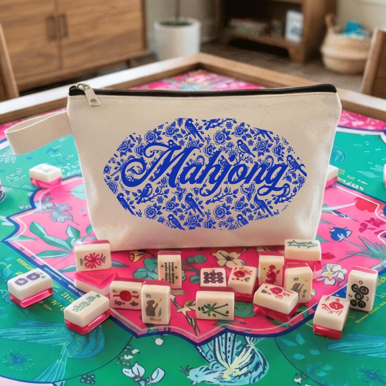 May include: A cream-colored zippered pouch with a blue floral and bird design and the word "Mahjong" in blue script. Several Mahjong tiles with red and white accents are scattered in front of the pouch, on a colorful patterned surface.