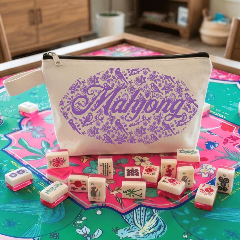 May include: A cream-colored zippered pouch with a purple floral and bird design that spells out "Mahjong". The pouch is on a colorful game board, surrounded by Mahjong tiles with red and white accents.
