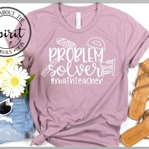 May include: A mauve t-shirt with the text "Problem Solver #mathteacher" in white. The text is surrounded by math-related icons, including a ruler, protractor, and an abacus.