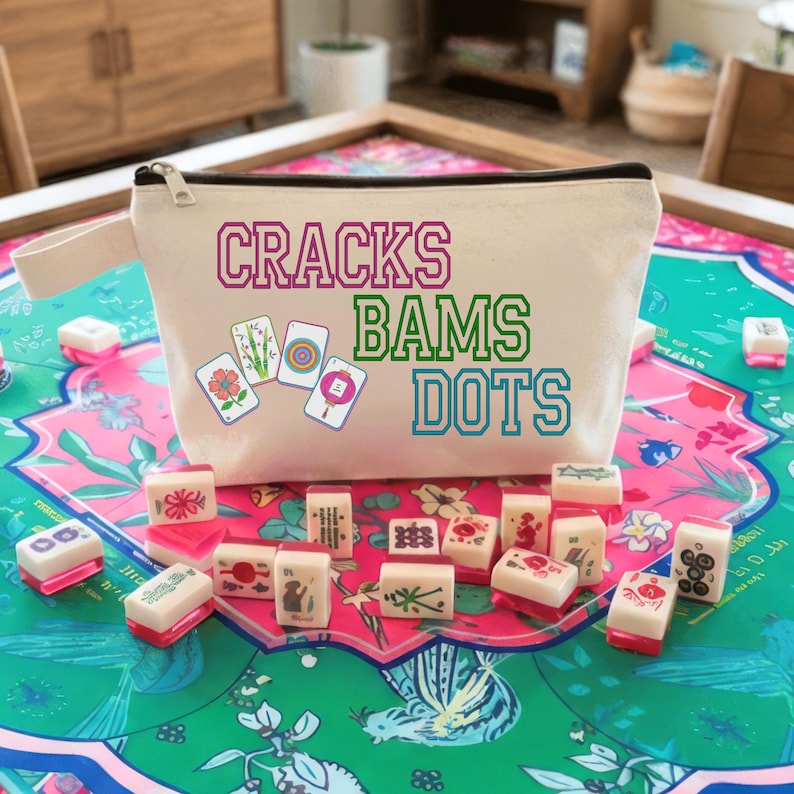 May include: A cream-coloured zipped pouch with the words "CRACKS BAMS DOTS" in pink, green, and blue. The pouch is surrounded by Mahjong tiles and sits on a colourful floral patterned surface. The pouch has a black zip.