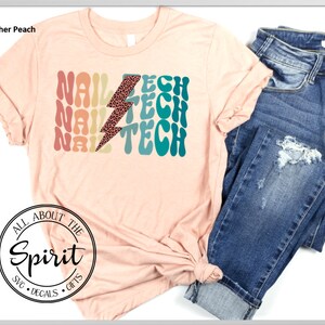 Op de afbeelding: Perzikkleurig T-shirt met de tekst "NAIL TECH" in retro-lettertype en een bliksemschicht met luipaardprint. Het shirt wordt gecombineerd met een blauwe jeans. De mouwen zijn opgerold en het shirt is in de taille geknoopt.