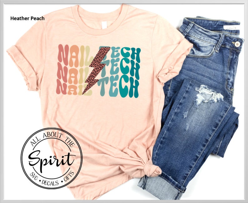 Op de afbeelding: Perzikkleurig T-shirt met de tekst "NAIL TECH" in retro-lettertype en een bliksemschicht met luipaardprint. Het shirt wordt gecombineerd met een blauwe jeans. De mouwen zijn opgerold en het shirt is in de taille geknoopt.