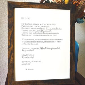 May include: A framed print of a poem titled "Hell Cat" about a daughter who is loud and opinionated. The poem reads: "My daughter screams with her whole body. Fists clenched, face red, belly tight. She hasn't learned words like demure, like obedient. There is no meekly, no softly, no quiet. There is only instinct and emotion and appetite. There is only need and persistence and fight. When she cries, she wants the whole world to hear it. When she opens her mouth, she doesn't care who's rattled by the sound. People say things like difficult, like opinionated, bossy and loud. Good, I tell her. Never be silent. Scream on, little hell cat, scream on. L.E. Bowman."