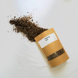 May include: A brown paper bag with the text "Potting Soil" printed on a white label. The bag is filled with dark brown potting soil and is spilling out onto a white surface.