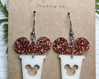 May include: A pair of white glitter coffee cup earrings with a Mickey Mouse silhouette cut out. The coffee cups are topped with brown glitter Mickey Mouse ears.