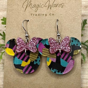May include: A pair of pink glitter bow earrings with a black and white patchwork design. The earrings are shaped like Mickey Mouse ears and have silver hooks.