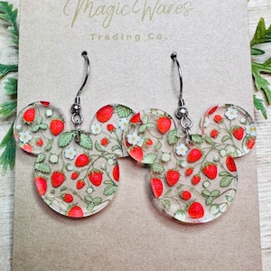 May include: A pair of silver dangle earrings with clear round acrylic charms featuring a strawberry pattern. The charms are shaped like Mickey Mouse ears.