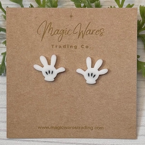 May include: A pair of white stud earrings shaped like Mickey Mouse's iconic white gloves with black detailing. The earrings are displayed on a brown card with the text "Magic Wares Trading Co." and "www.magicwarestrading.com".