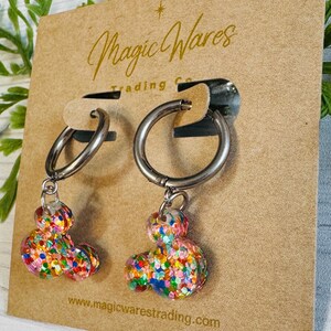 May include: A pair of silver hoop earrings with colourful confetti Mickey Mouse charms hanging from them. The earrings are displayed on a brown card with the text "Magic Wares Trading Co. www.magicwarestrading.com"