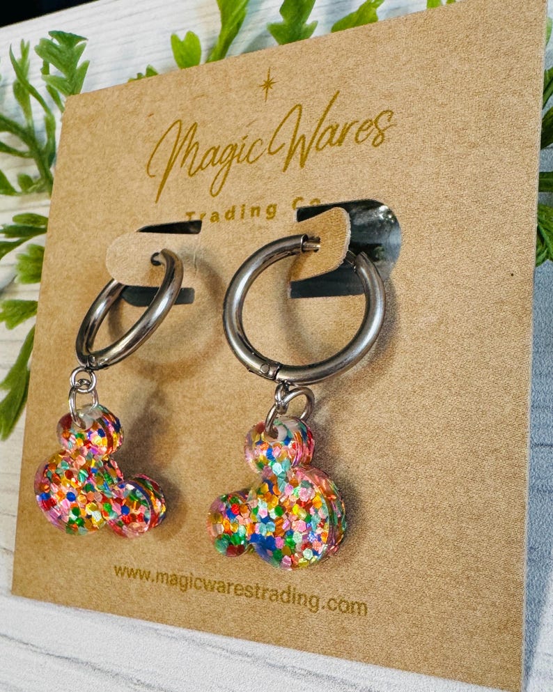 May include: A pair of silver hoop earrings with colourful confetti Mickey Mouse charms hanging from them. The earrings are displayed on a brown card with the text "Magic Wares Trading Co. www.magicwarestrading.com"
