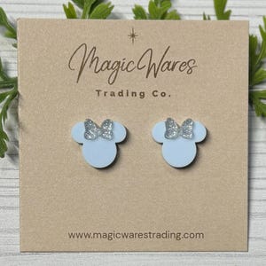May include: A pair of light blue acrylic earrings shaped like a mouse head with a silver glitter bow. The earrings are displayed on a tan card with the text "Magic Wares Trading Co." and the website address.