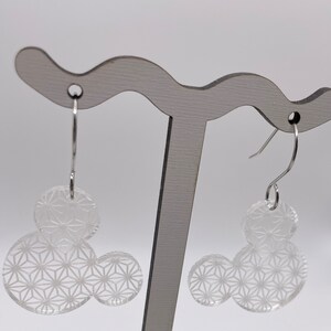 May include: A pair of silver dangle earrings with clear acrylic geometric shapes. The shapes are in the form of a Mickey Mouse head with a repeating pattern.