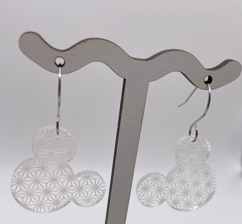 May include: A pair of silver dangle earrings with clear acrylic geometric shapes. The shapes are in the form of a Mickey Mouse head with a repeating pattern.