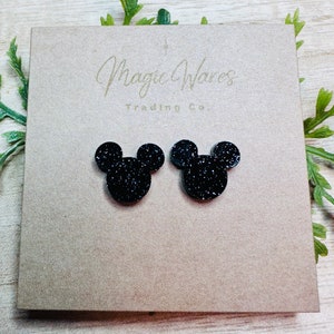 May include: A pair of black glitter Mickey Mouse earrings. The earrings are on a brown card with the text "Magic Wares Trading Co."