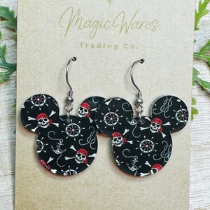 May include: A pair of black and white Mickey Mouse-shaped earrings with a pirate skull and crossbones pattern. The earrings have silver hooks and are displayed on a brown card with the text "Magic Wares Trading Co."