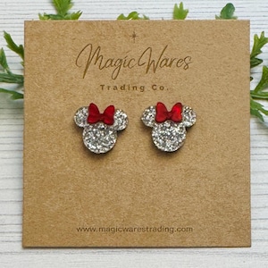May include: A pair of glittery silver mouse-shaped earrings with red bows, displayed on a brown card. The card has the text "Magic Wares Trading Co." and the website address www.magicwarestrading.com.