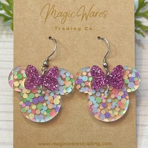 May include: A pair of clear acrylic earrings shaped like Mickey Mouse ears with a pink glitter bow and colorful confetti inside. The earrings are on a brown card with the text "Magic Wares Trading Co." and "www.magicwarestrading.com".