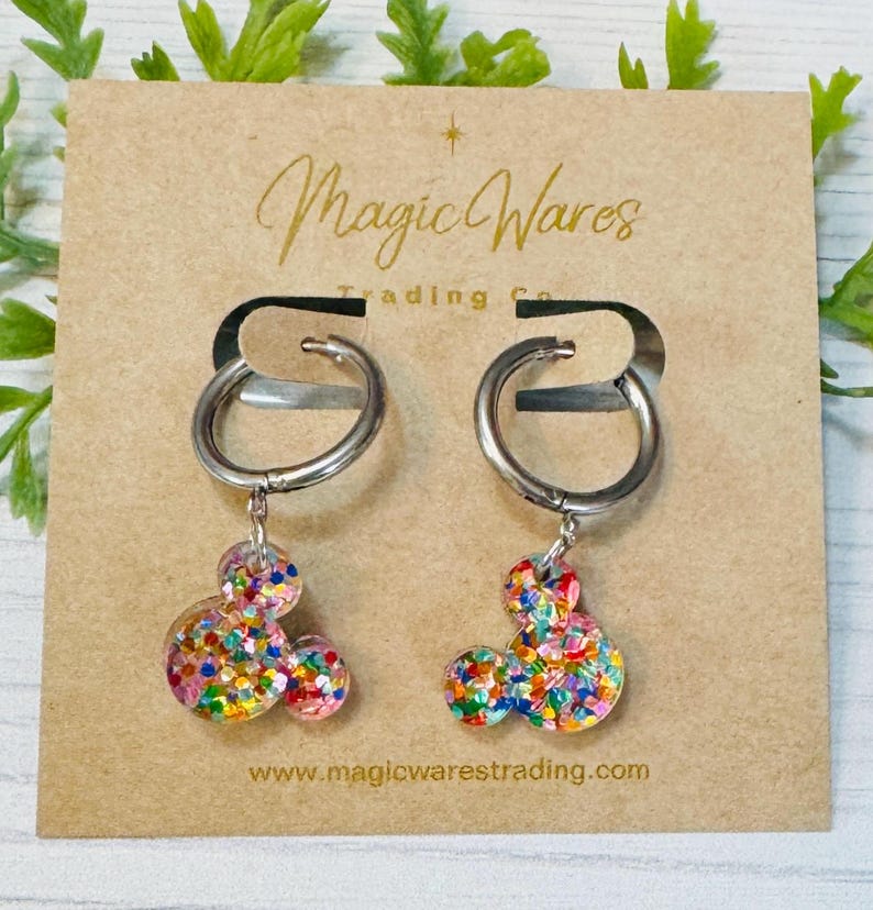 May include: A pair of silver hoop earrings with colourful confetti Mickey Mouse charms. The earrings are on a brown card with the text "Magic Wares Trading Co." and "www.magicwarestrading.com".
