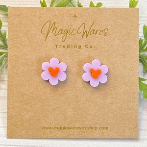 May include: A pair of purple flower-shaped stud earrings with orange Mickey Mouse heads in the center. The earrings are displayed on a brown card with the text "Magic Wares Trading Co." and "www.magicwarestrading.com".