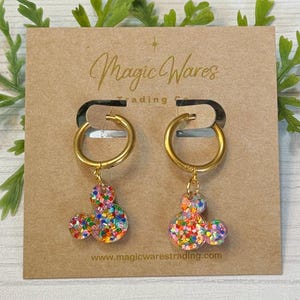 May include: Gold hoop earrings with a dangling Mickey Mouse charm. The charm is made of clear resin with colourful glitter embedded in it. The earrings are displayed on a brown card with the text "Magic Wares Trading Co. www.magicwarestrading.com".