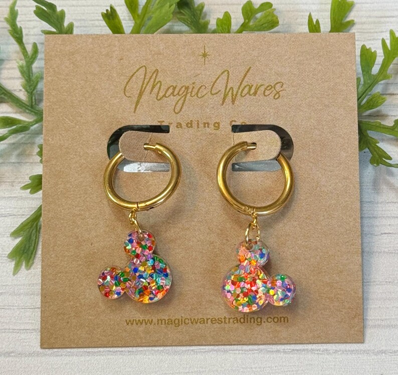 May include: Gold hoop earrings with a dangling Mickey Mouse charm. The charm is made of clear resin with colourful glitter embedded in it. The earrings are displayed on a brown card with the text "Magic Wares Trading Co. www.magicwarestrading.com".