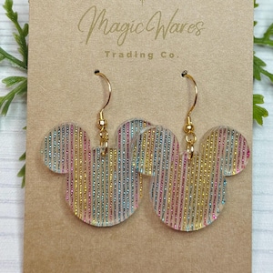 May include: Earrings featuring a Mickey Mouse head design with vertical stripes in rainbow colors. The earrings are attached to gold-colored hooks and are displayed on a tan card with the text "Magic Wares Trading Co."