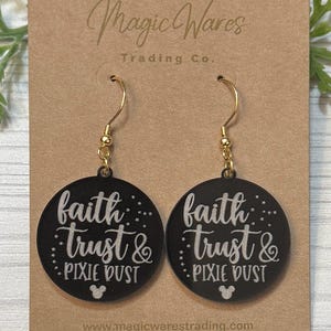 May include: Black round earrings with gold-colored hooks. Each earring features the words "faith, trust & pixie dust" in white script, with a small Mickey Mouse silhouette at the bottom. The earrings are displayed on a brown card.