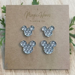 May include: Four gray and white Mickey Mouse-shaped earrings with a geometric pattern. The earrings are displayed on a brown card with the text "Magic Wares Trading Co."