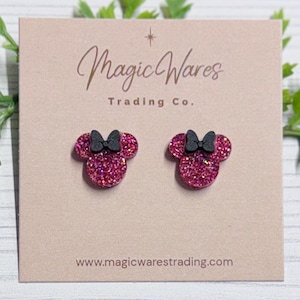 May include: A pair of pink glitter Minnie Mouse-shaped stud earrings with black bows, displayed on a pink card. The card has the text "Magic Wares Trading Co." and the website address www.magicwarestrading.com.