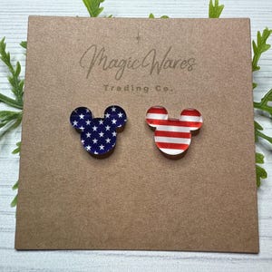 May include: Two pairs of Mickey Mouse-shaped earrings with an American flag design. One pair features red and white stripes, and the other pair features blue stars on a white background. The earrings are displayed on a brown card with the text "Magic Wares Trading Co."