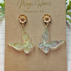 May include: A pair of gold-colored earrings with a butterfly design. The earrings have a flower-shaped top and a butterfly-shaped pendant with intricate cut-out details. The earrings are displayed on a brown card with the text "Magic Wares Trading Co."