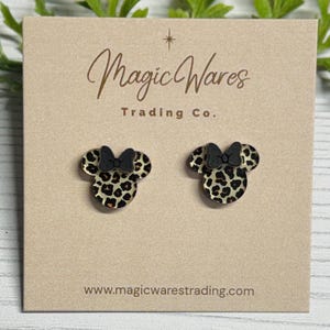 May include: A pair of stud earrings shaped like a mouse head with a black bow. The earrings have a leopard print pattern. The earrings are displayed on a tan card with the text "Magic Wares Trading Co." and the website address.