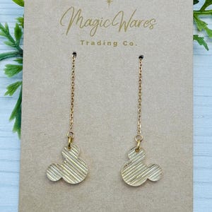 May include: A pair of gold dangle earrings with a Mickey Mouse head design. The earrings are on a card that says "Magic Wares Trading Co."