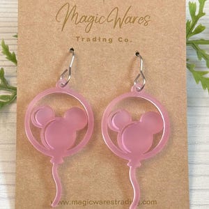 May include: Pink acrylic earrings shaped like Mickey Mouse balloons. The earrings are on a brown card with the text "Magic Wares Trading Co." and "www.magicwarestrading.com".