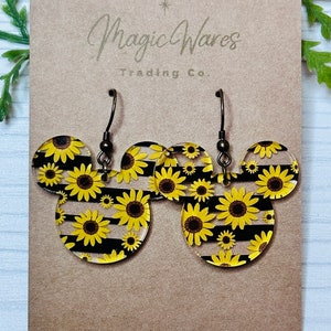 May include: A pair of dangle earrings with a black and white striped background and a sunflower pattern. The earrings are made of clear acrylic and have a silver hook.