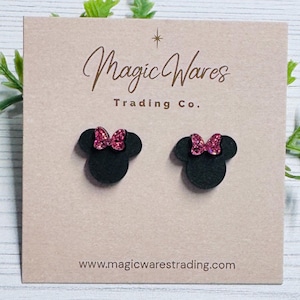 May include: A pair of black mouse-shaped stud earrings with pink glitter bows, displayed on a light brown card. The card features the text "Magic Wares Trading Co." and the website address www.magicwarestrading.com.