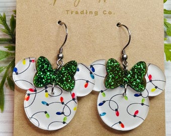 May include: A pair of white acrylic earrings shaped like Mickey Mouse ears with a green glitter bow and a string of colorful lights printed on them. The earrings are on a brown card with the text "Magic Wares Trading Co."
