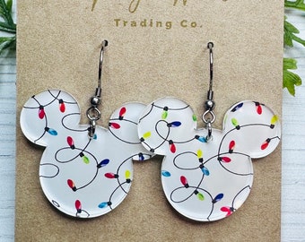May include: A pair of clear acrylic earrings in the shape of a mouse head, featuring a design of colorful Christmas lights. The earrings hang from silver-colored ear wires and are displayed on a brown card with the text "Magic Wares Trading Co."