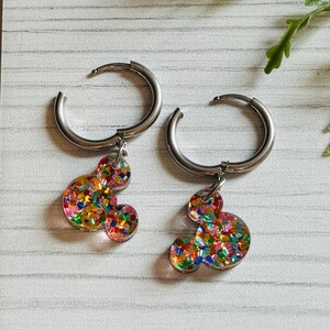May include: A pair of silver hoop earrings with colourful glitter Mickey Mouse charms. The charms are filled with red, orange, yellow, green, blue, and purple glitter.