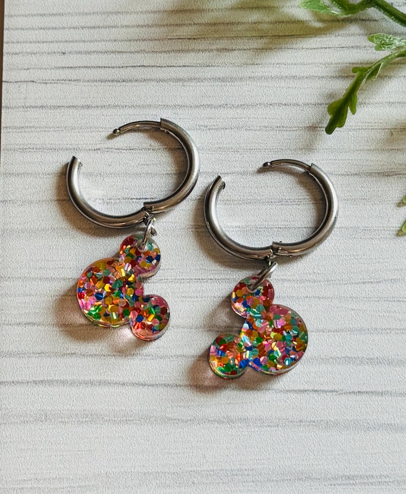 May include: A pair of silver hoop earrings with colourful glitter Mickey Mouse charms. The charms are filled with red, orange, yellow, green, blue, and purple glitter.