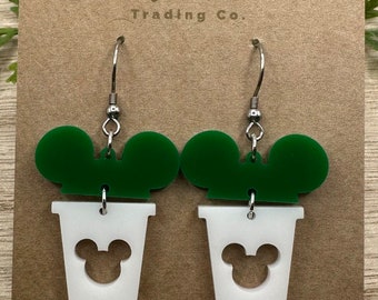 May include: A pair of green and white dangle earrings featuring a Mickey Mouse silhouette. The earrings are made of acrylic and have silver hooks.