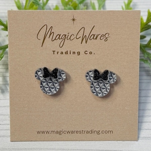 May include: A pair of stud earrings shaped like Minnie Mouse heads. The earrings have a geometric pattern in gray and white, with a black bow. The earrings are displayed on a tan card with the text "Magic Wares Trading Co." and the website address.