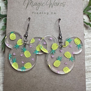 May include: Clear acrylic earrings shaped like Mickey Mouse ears with a pineapple print and pink hearts. The earrings are on a brown card with the text "Magic Wares Trading Co."